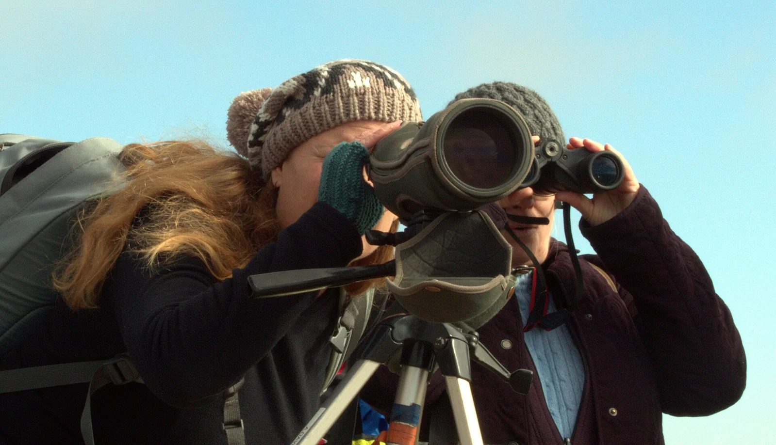 Two observers using a spotting scope and binoculars against open sky.
