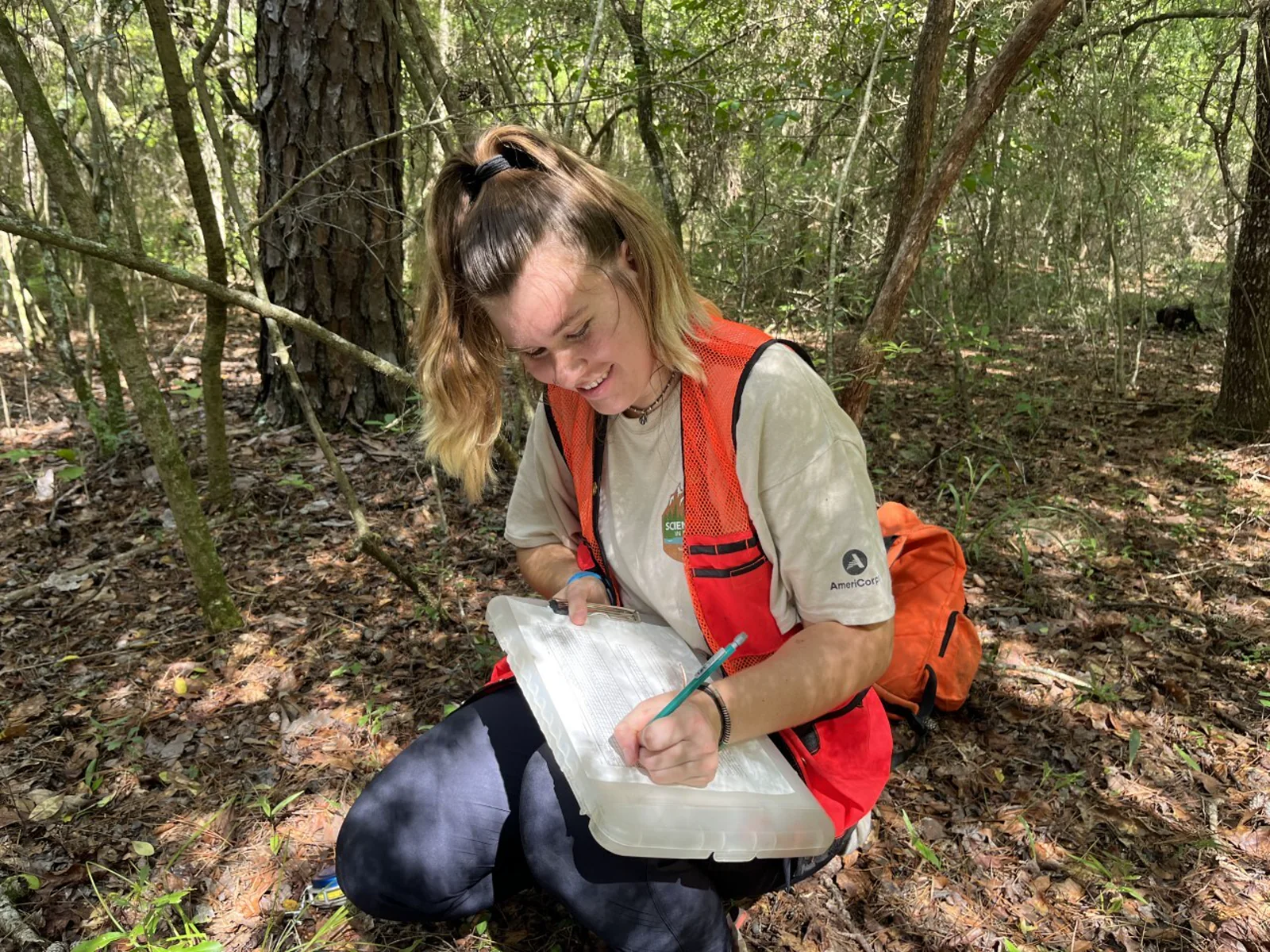 A field scientist kneeling among trees and recording observations on a clipboard.