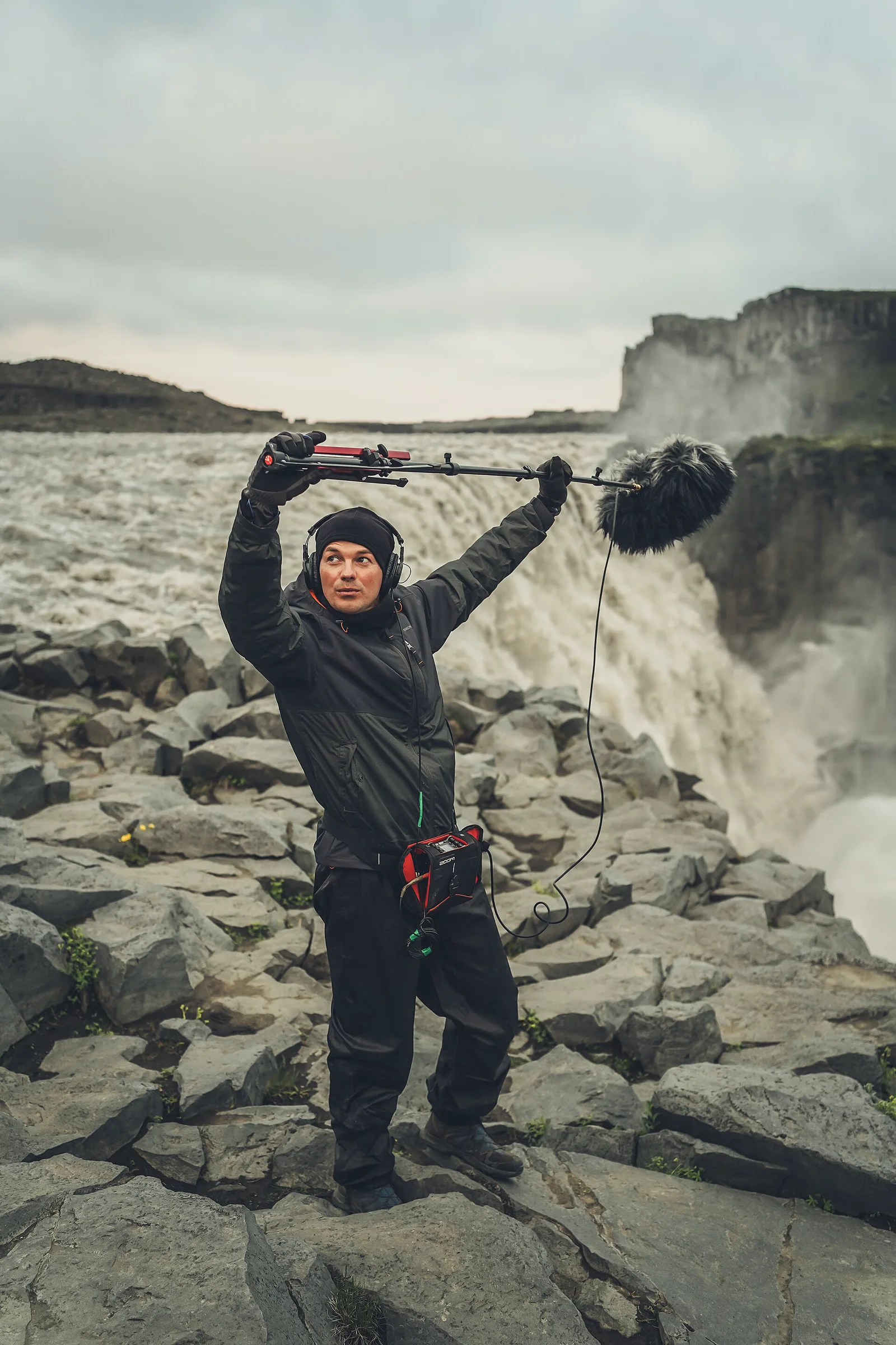 A field recordist wearing headphones and holding a wind-protected microphone outdoors.